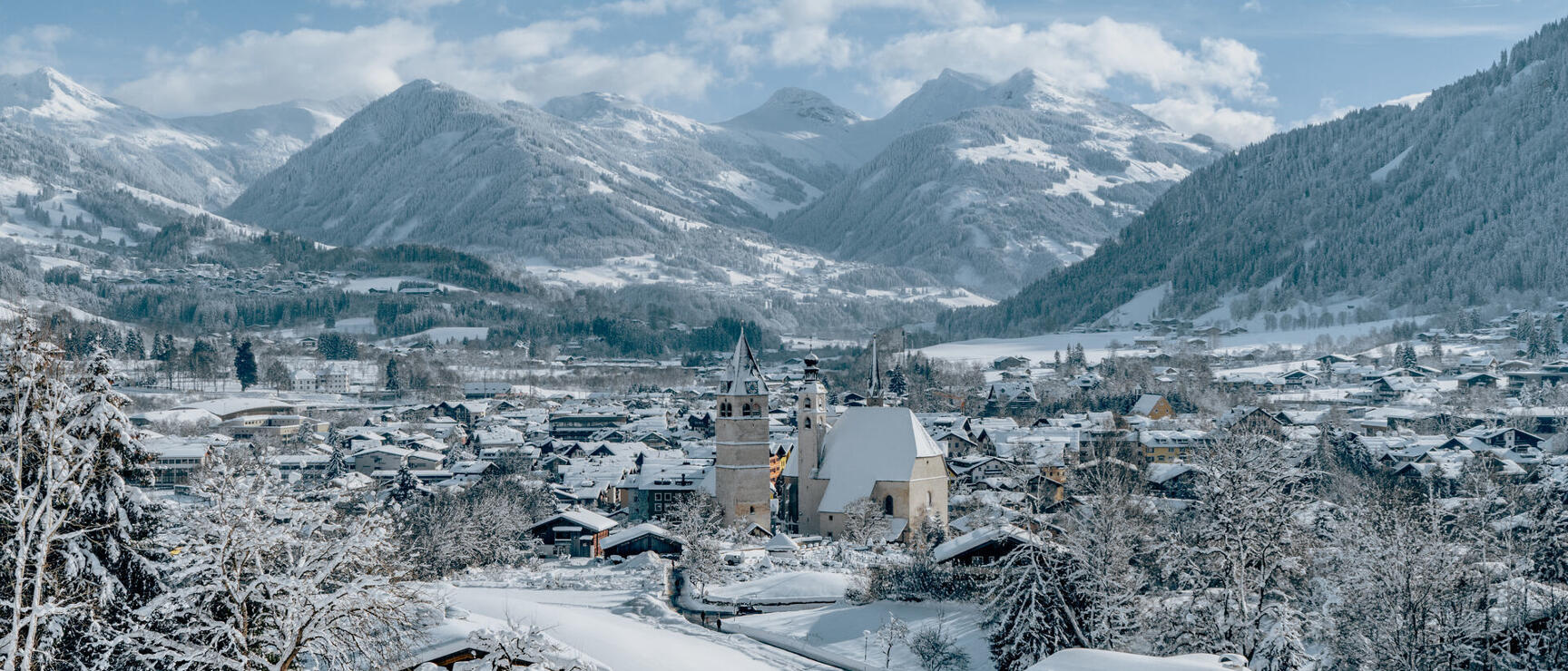 Snowy village with church tower in the valley, snow-covered mountains and forests in the background.
