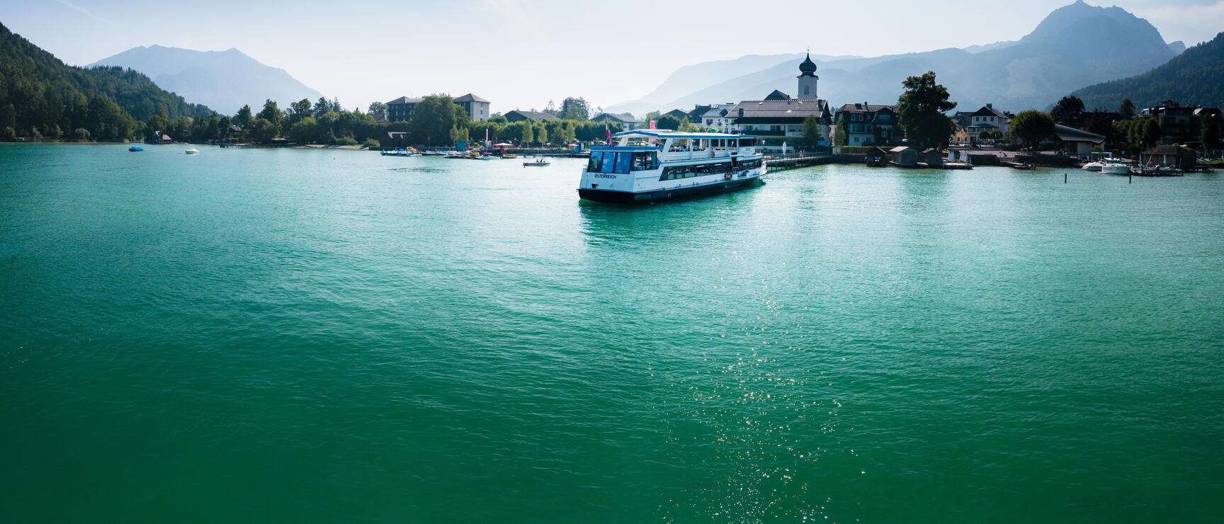 White passenger ship on turquoise lake, village with church tower and mountain panorama in the background.