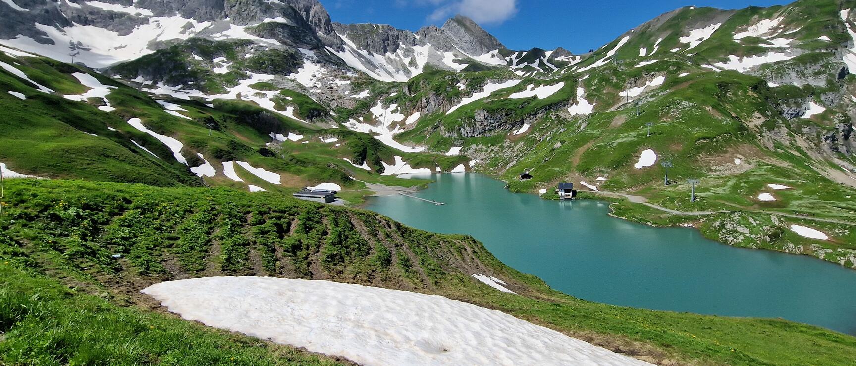 A view from above of Lake Zürs, nestled between mountains and lush green meadows, still partly covered in snow