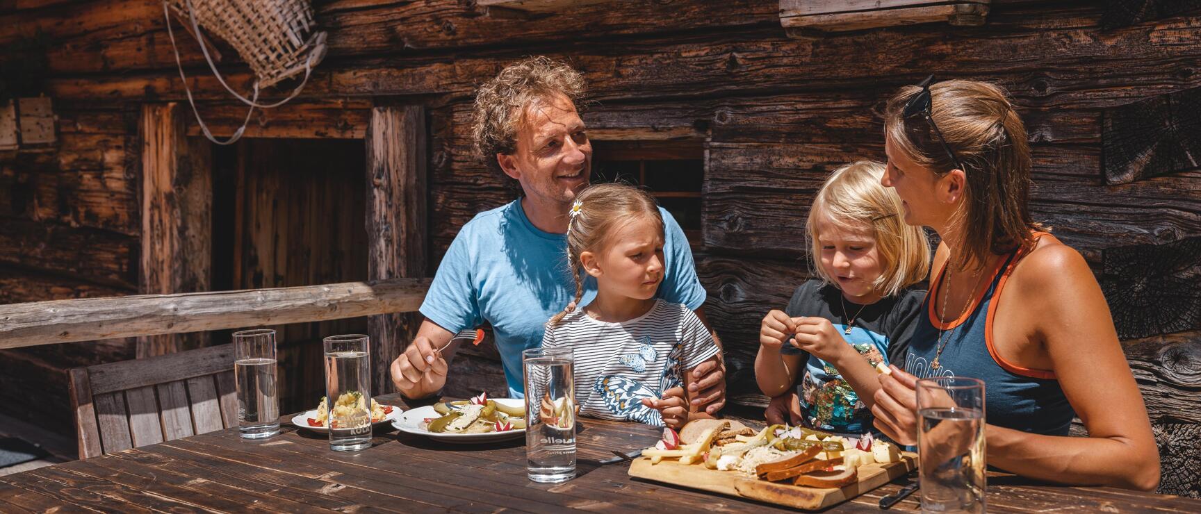 Two adults and two children eating at a wooden table in front of a cabin at the Fahrmkehralm in Inneralpbach; a Brettljause on the table