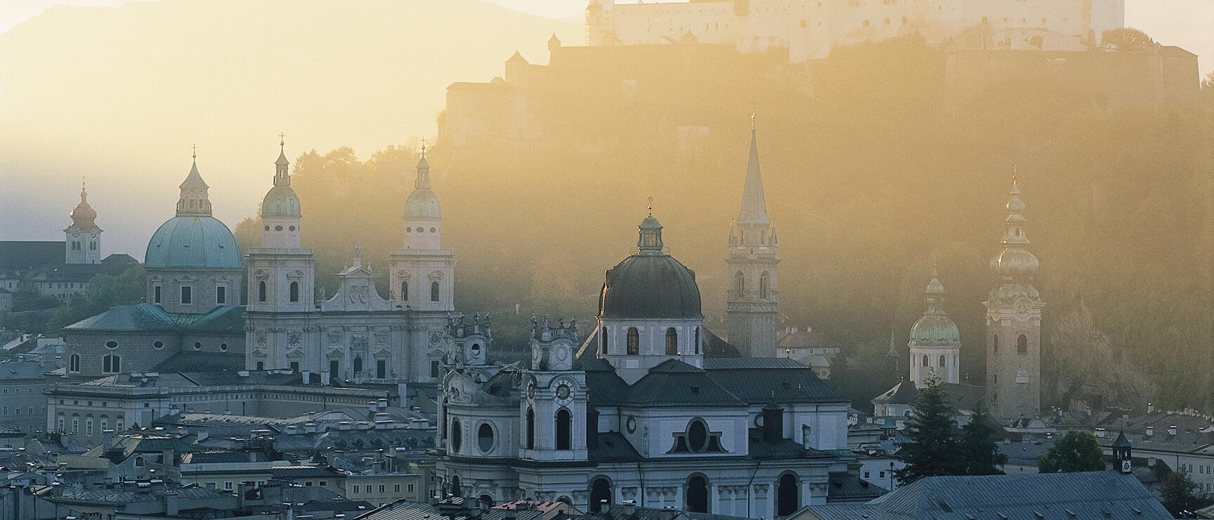 Panoramic view of Salzburg's Old Town with church domes and Hohensalzburg Fortress in morning mist.