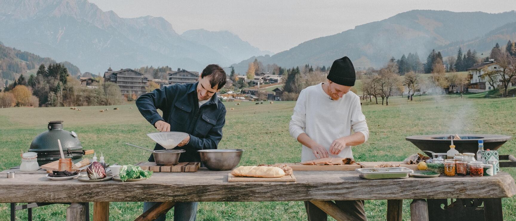 Two people cooking at a wooden table outdoors, ingredients and grills, Alpine panorama in the background.