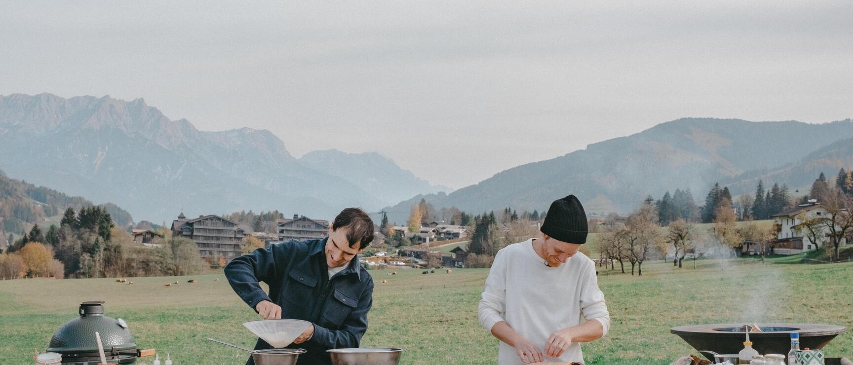 Two people cooking at a wooden table outdoors, ingredients and grills, Alpine panorama in the background.