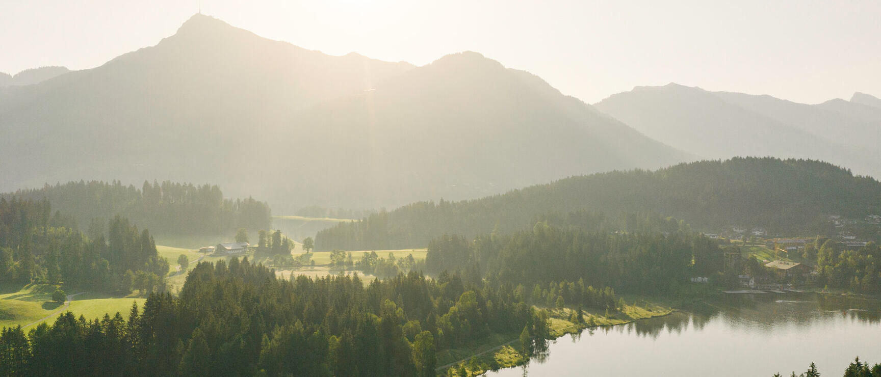 Aerial view of a lake surrounded by coniferous forests, mountain panorama backlit by the sun.