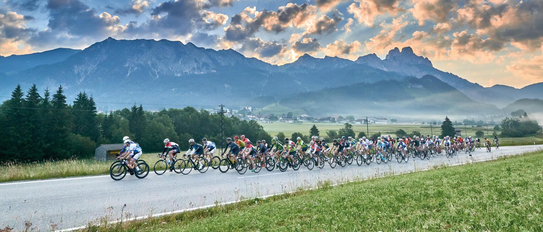 Large group of cyclists on a country road with mountain panorama and dramatic cloudy sky in the background.