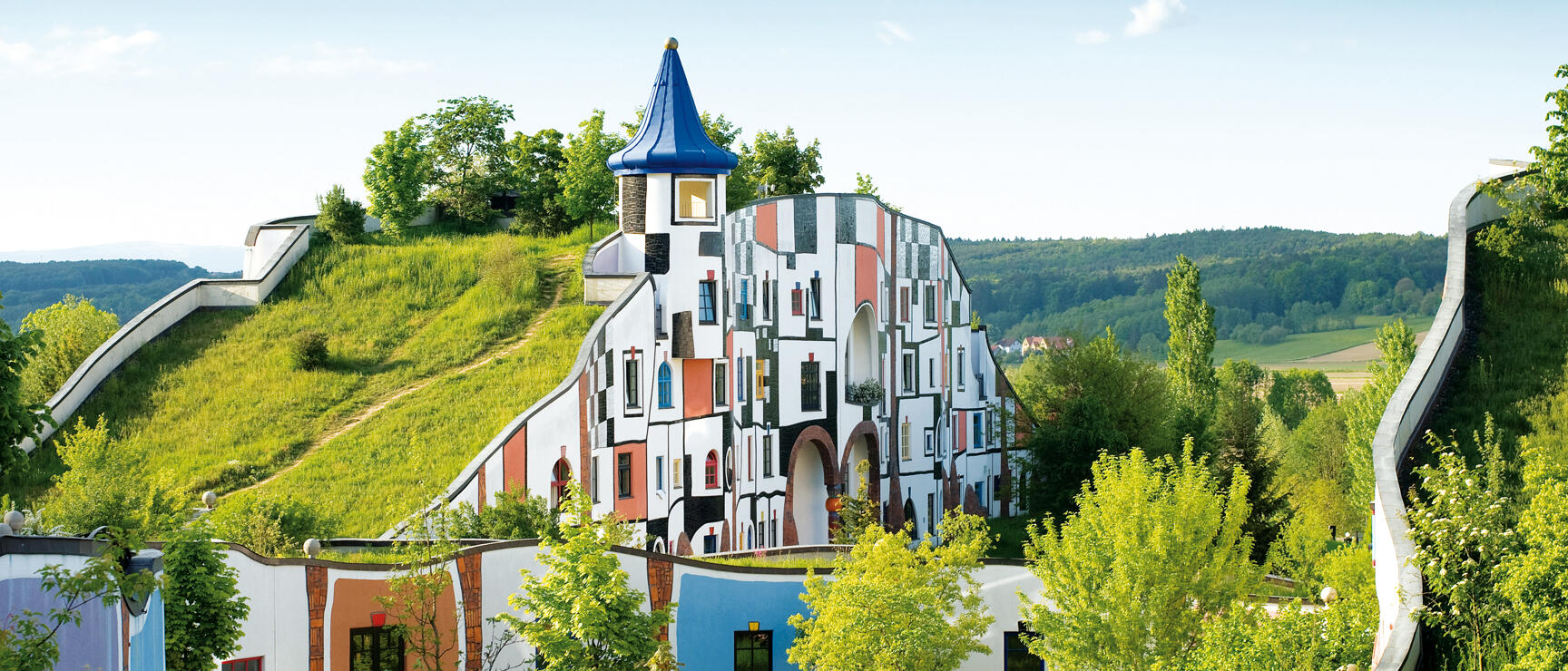 Rogner Bad Blumau: colourful Hundertwasser building with grass roof, blue tower and curved façade, hills in background.