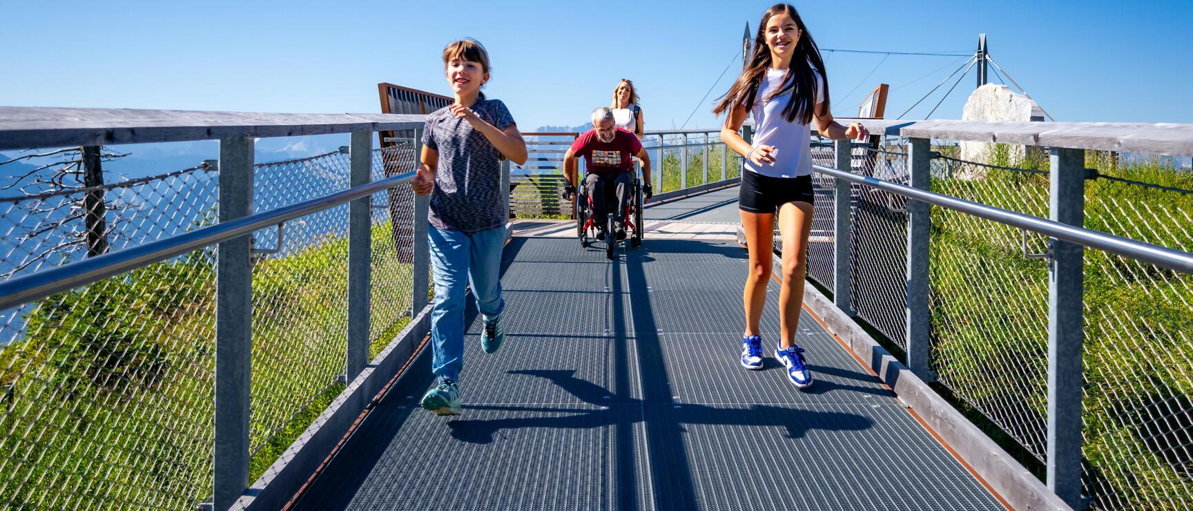 Two children running on a metal footbridge, wheelchair user and woman behind them, blue sky.
