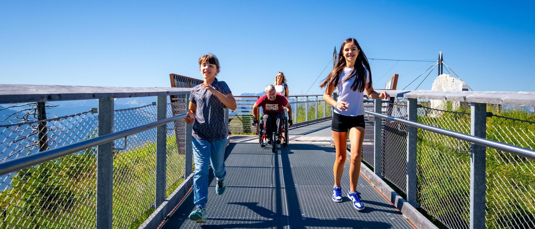 Two children running on a metal footbridge, wheelchair user and woman behind them, blue sky.