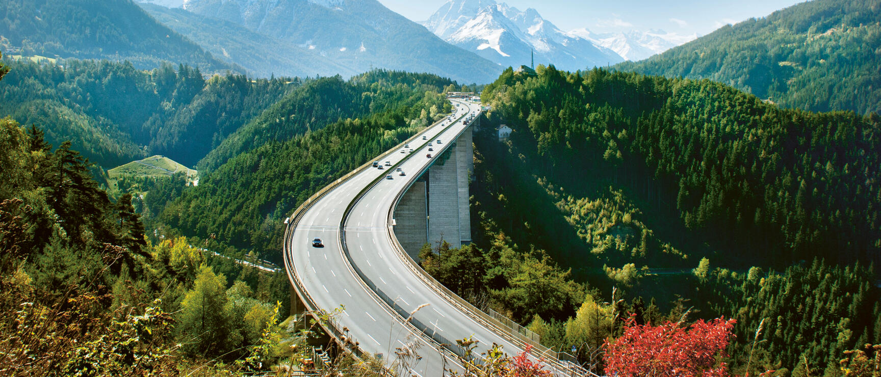 Autobahnbrücke durch bewaldete Alpenlandschaft, schneebedeckte Gipfel im Hintergrund, blauer Himmel.