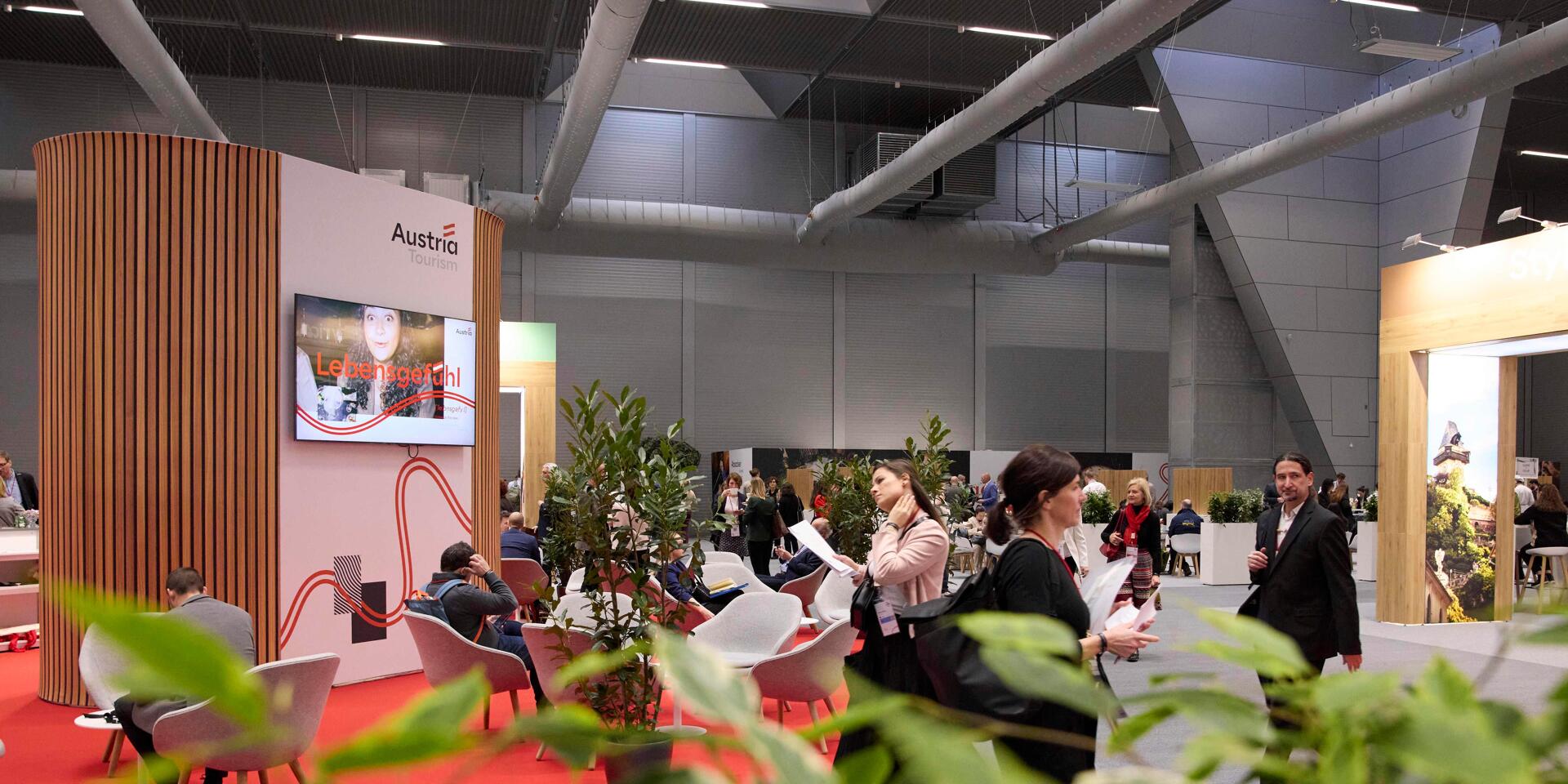 Austria Tourism trade fair stand with seating area, screen and plants, several people in an exhibition hall.