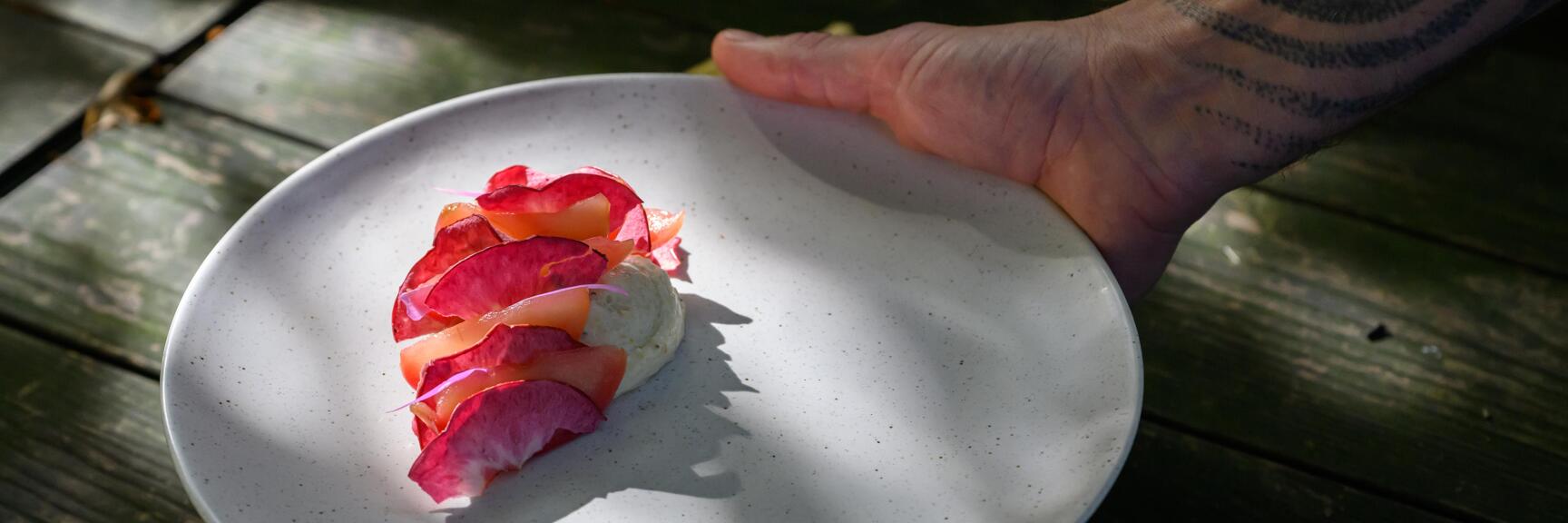 Riebelmais-Mousse mit dünnen rosa-orangen Scheiben und einem violetten Blütenfaden garniert, auf einem Teller, der von einer Hand im Freien gehalten wird.