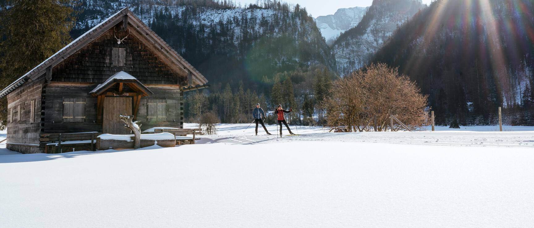 Two people cross-country skiing next to a wooden hut, snow-covered mountain landscape and sunrays in the background.