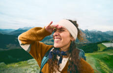 Smiling woman with headband and backpack shielding her eyes, mountain panorama in background.