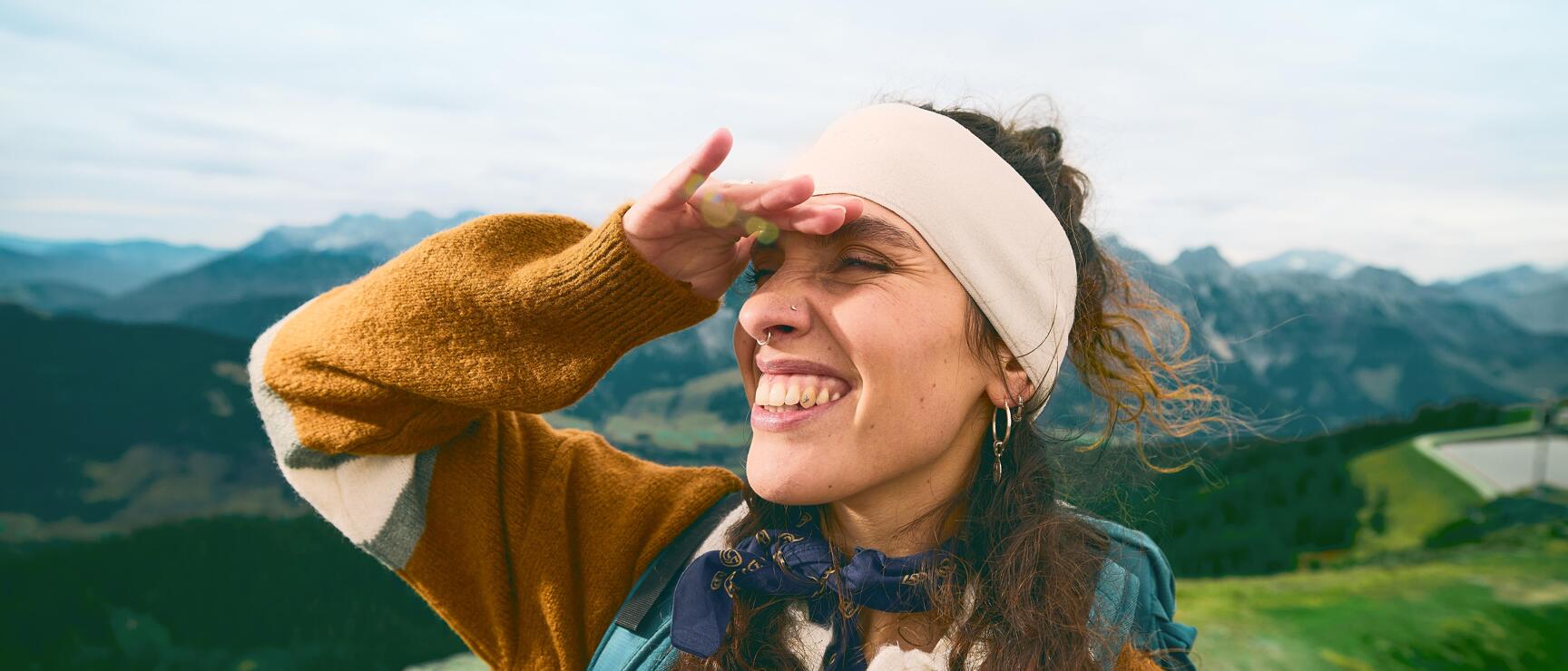 Smiling woman with headband and backpack shielding her eyes, mountain panorama in background.