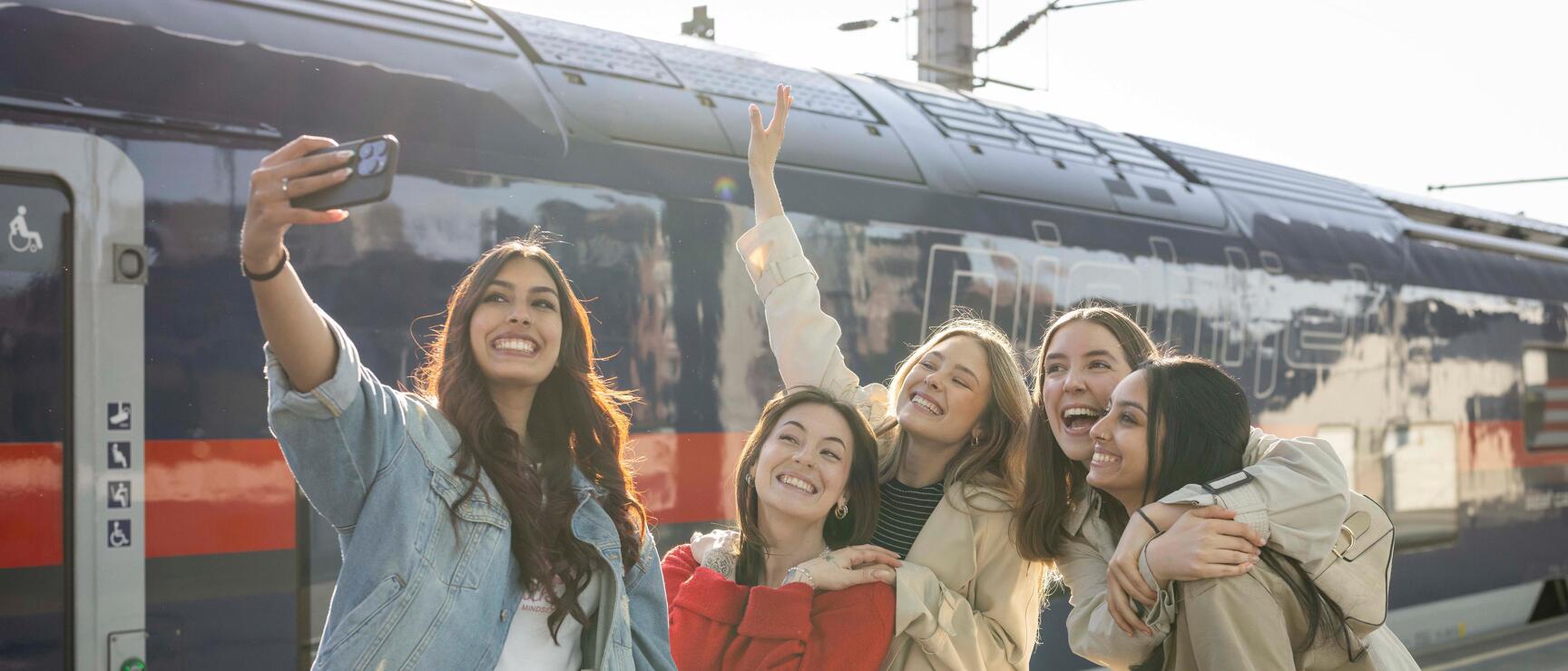 Five young women taking a selfie on a platform in front of an ÖBB Railjet train with luggage.