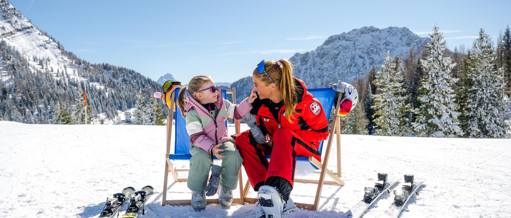 Ski instructor in red suit and child in deckchairs in snow, skis beside them, snow-covered mountains in background.