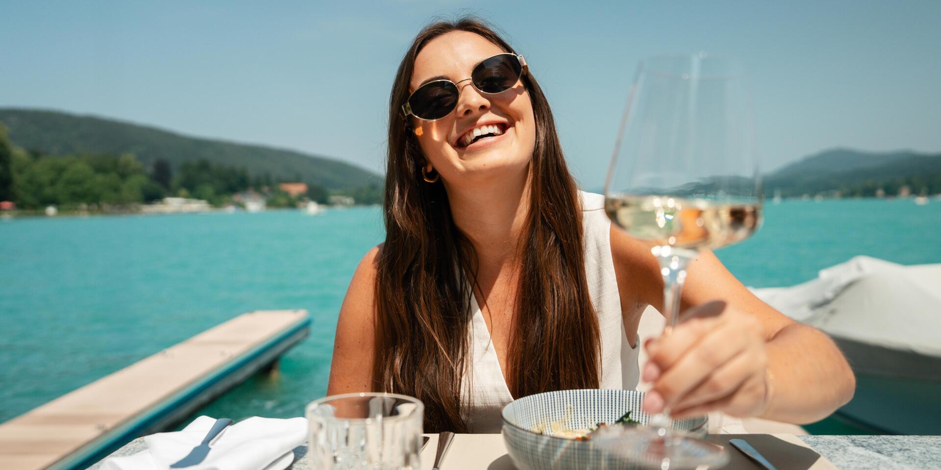 Woman wearing sunglasses holding a wine glass at a lakeside terrace table, turquoise lake and hills in the background.