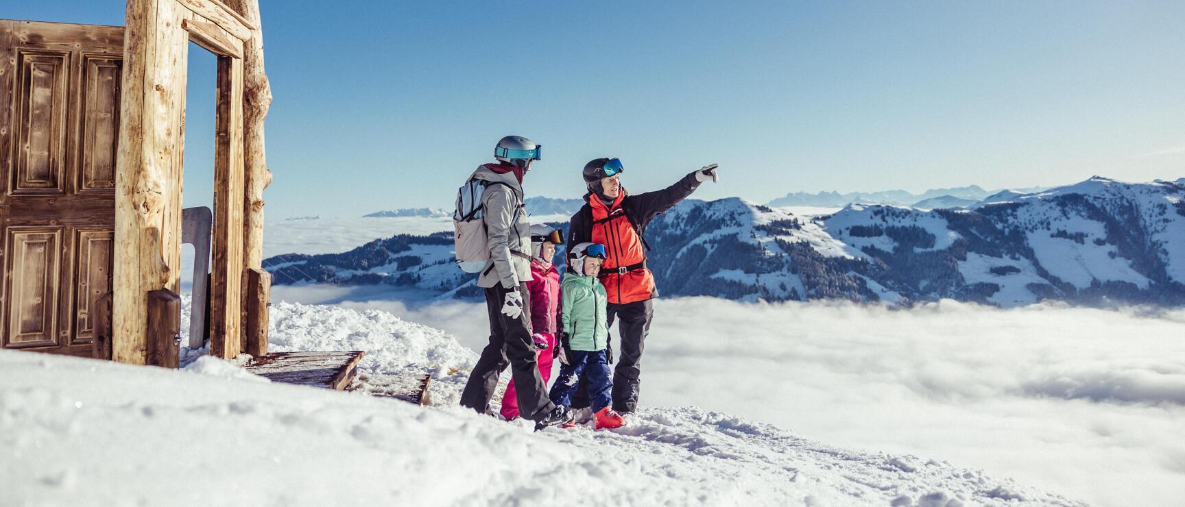 Four people in ski gear next to a wooden hut on a snow-covered summit with mountain panorama above clouds.