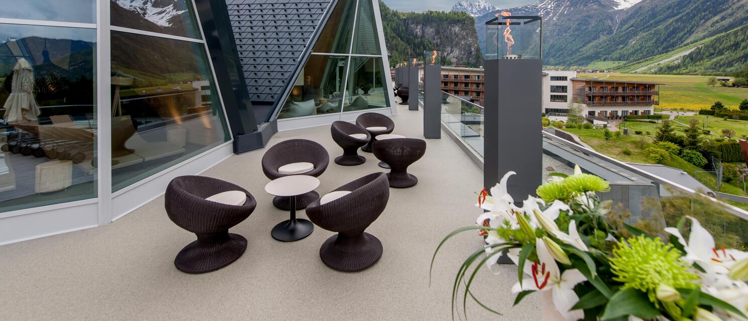 Hotel terrace with dark lounge chairs, fire columns and snowy mountains in the background.