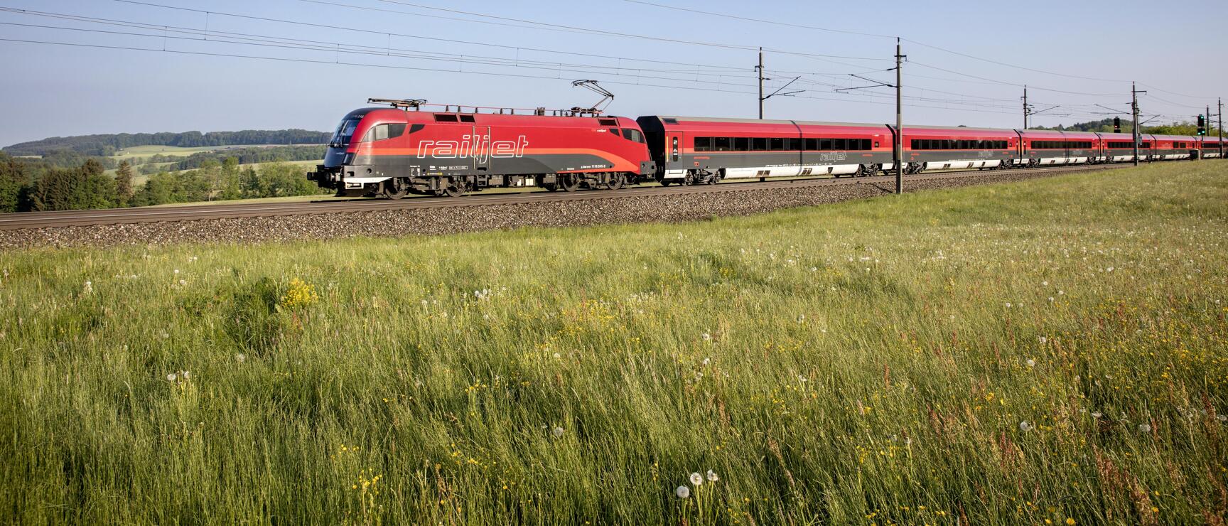 Red Railjet train travelling through green summer meadow under blue sky.