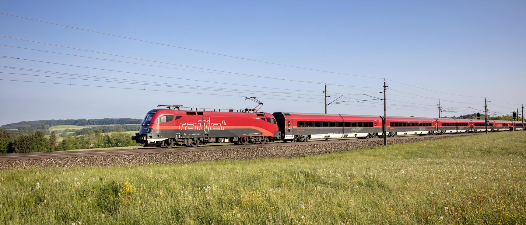 Red Railjet train travelling through green summer meadow under blue sky.