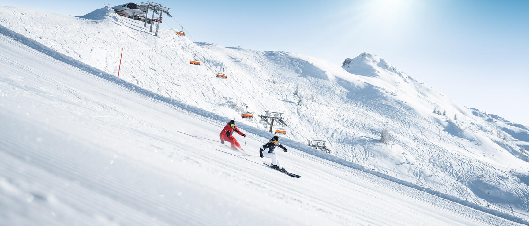 Two skiers on sunny slope, orange chairlift and mountain station in background.