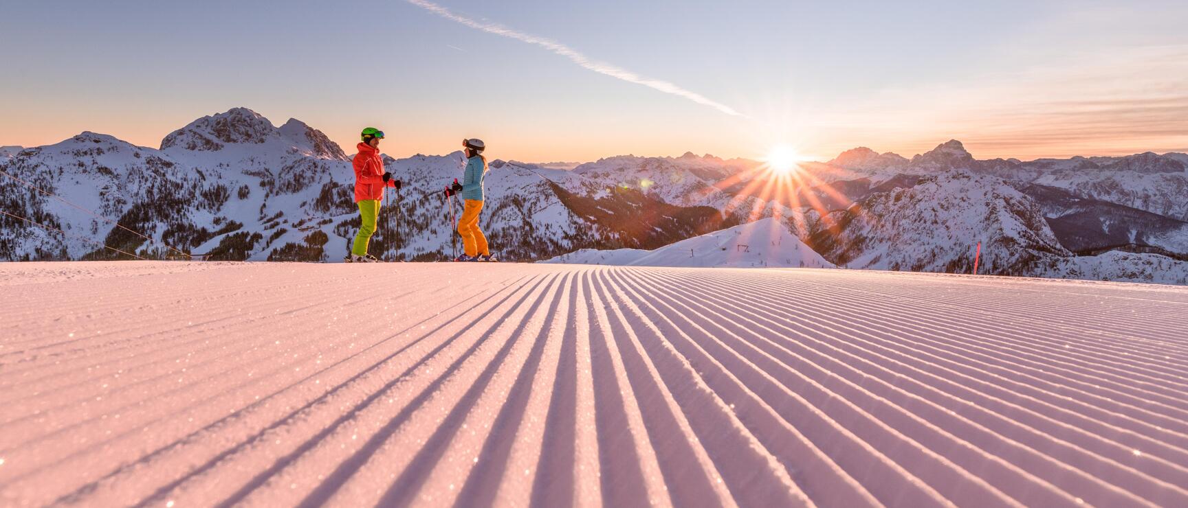 Two skiers on a groomed slope at sunrise with snow-covered mountain peaks in the background.
