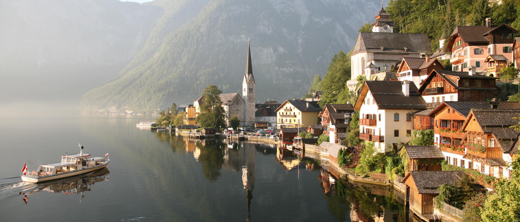 Hallstatt on Hallstatt Lake with church, wooden houses, a boat on the lake and mountains in morning mist.