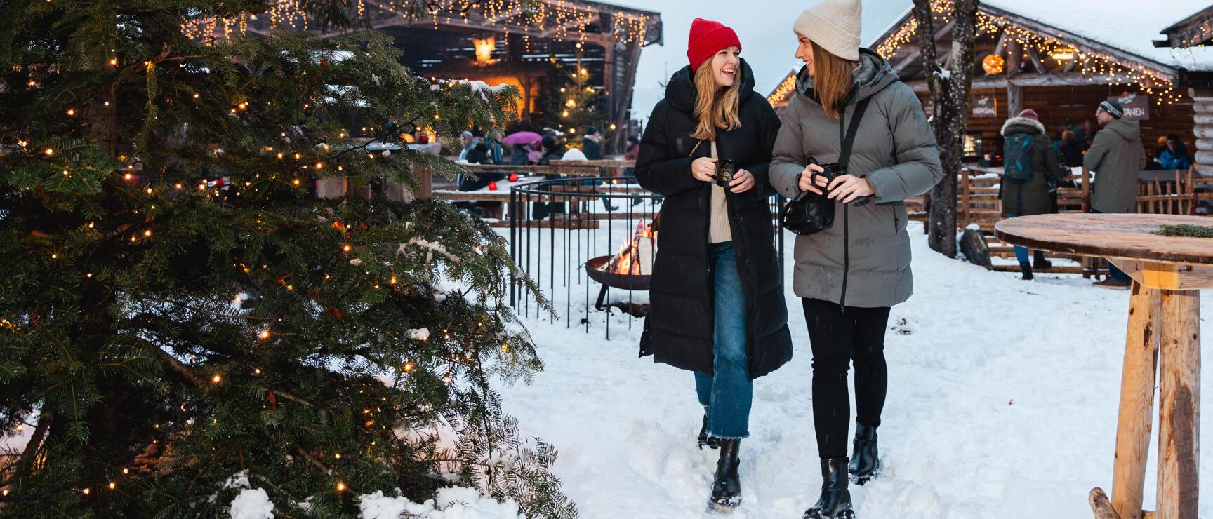 Two women wearing hats at snowy Christmas market, lit wooden huts and evergreen tree in snow.