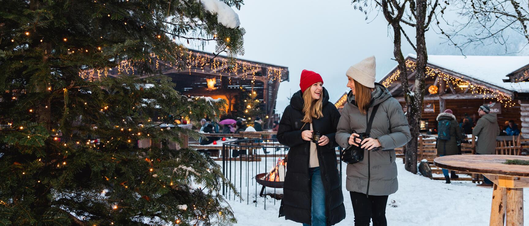 Zwei Frauen mit Mützen auf winterlichem Weihnachtsmarkt, beleuchtete Holzhütten und Tannenbaum im Schnee.