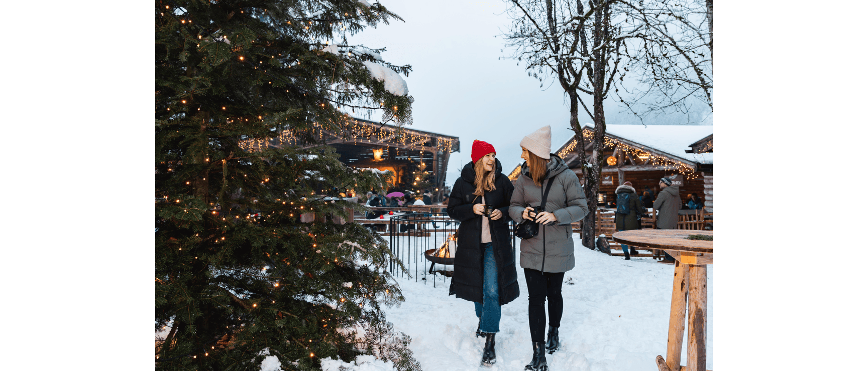 Two women wearing hats at snowy Christmas market, lit wooden huts and evergreen tree in snow.