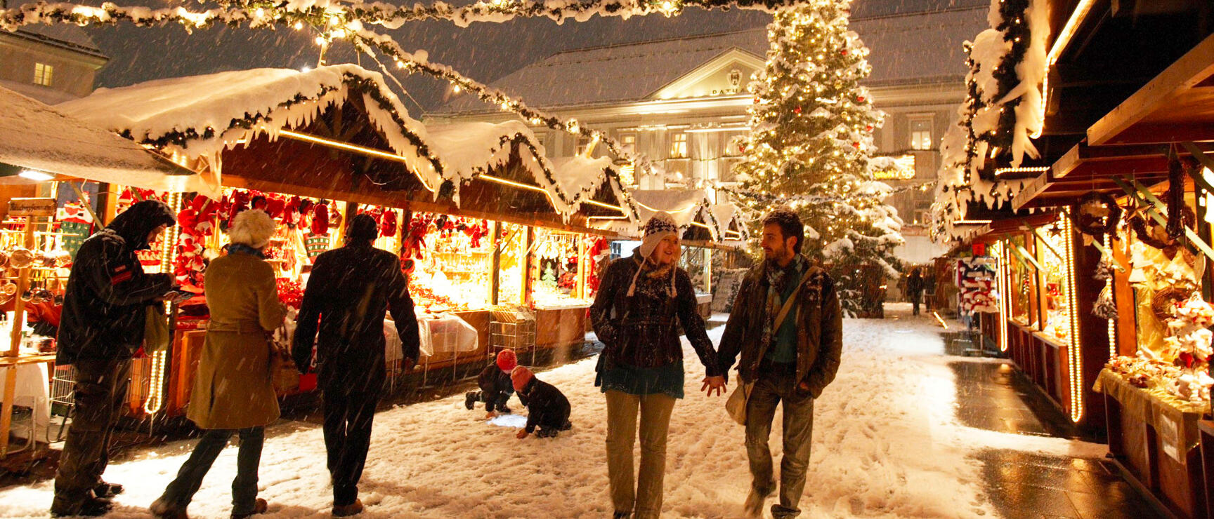 Christmas market during snowfall with illuminated wooden stalls, Christmas tree, and people strolling between stands