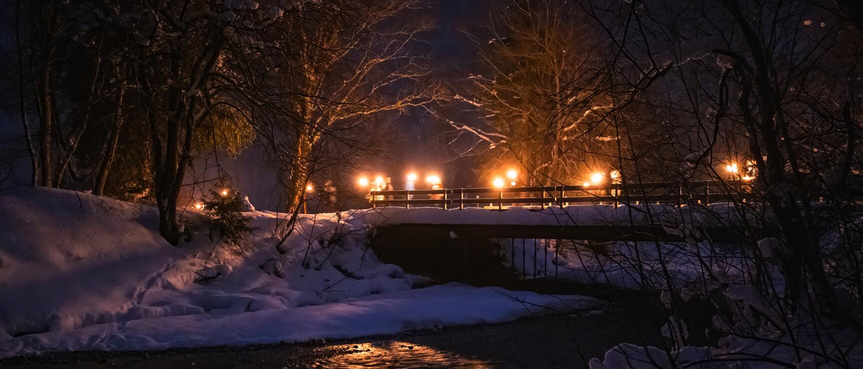 Snowy bridge with fire bowls at night, light reflection in stream, bare trees.