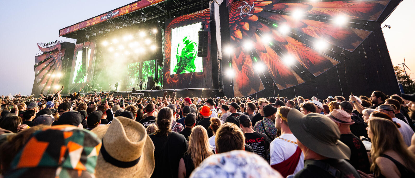 Large festival stage with green LED screen and wing artwork at Nova Rock, dense crowd in front.