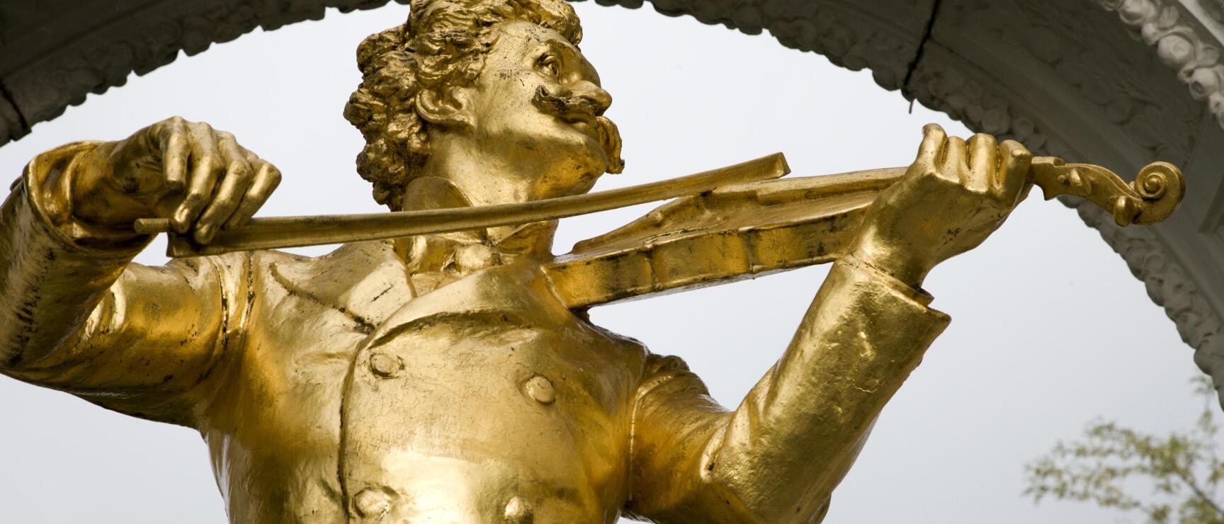 Golden Johann Strauss statue with violin in Vienna City Park, white marble arch in background.