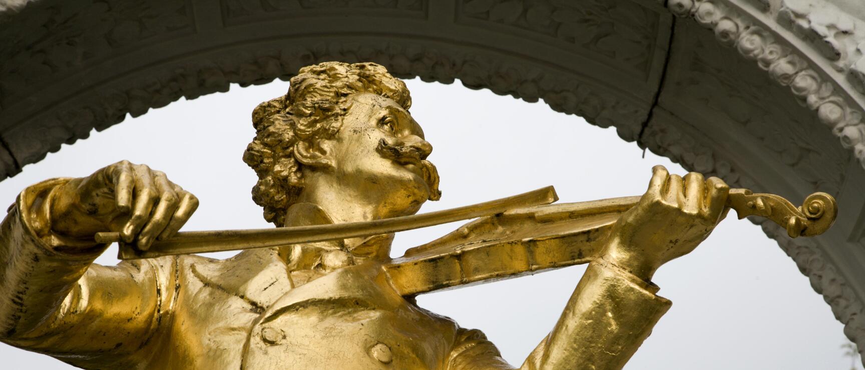 Goldene Johann-Strauss-Statue mit Geige im Stadtpark Wien, weißer Marmorbogen im Hintergrund.