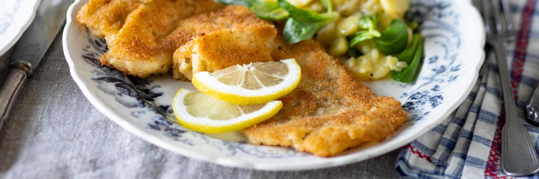 Wiener schnitzel with lemon slices and potato salad on blue and white plate, two white wine glasses in background.