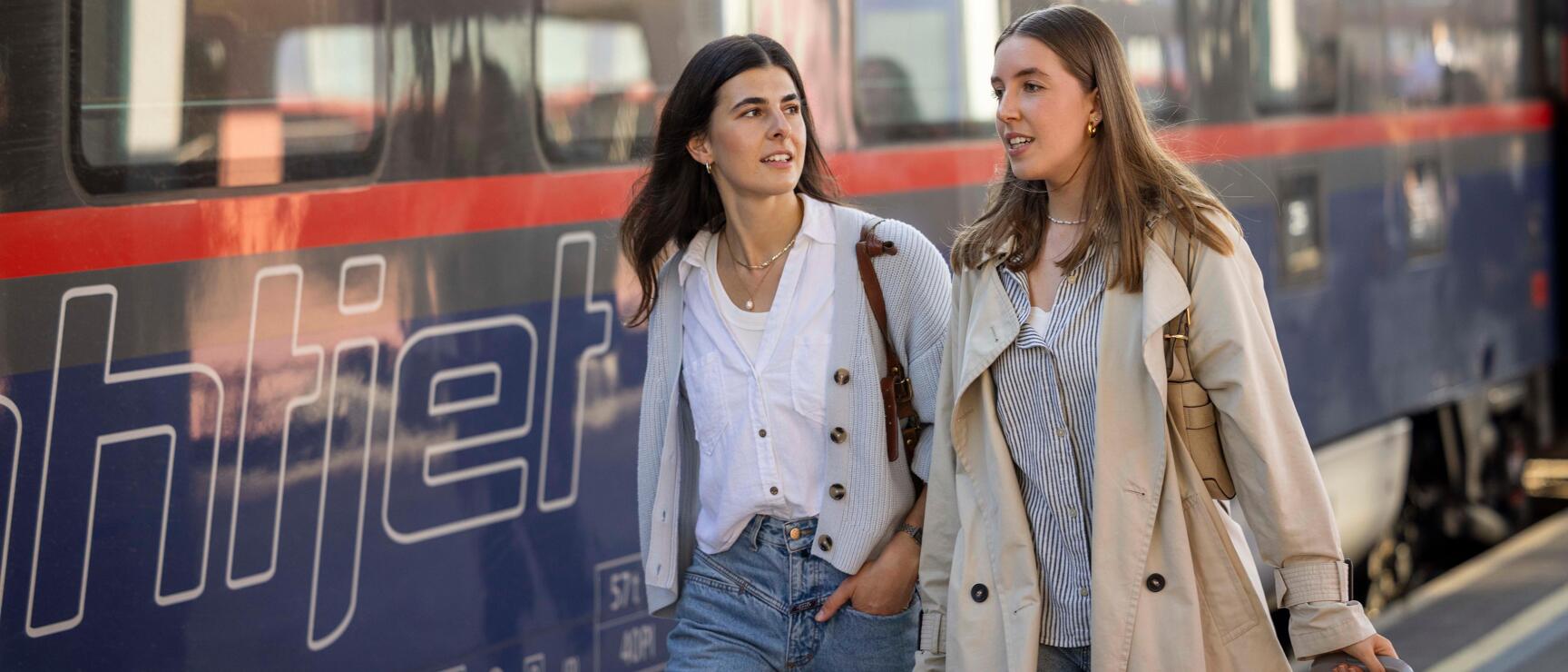 Two young women with suitcase walking along an ÖBB Railjet train on the platform.