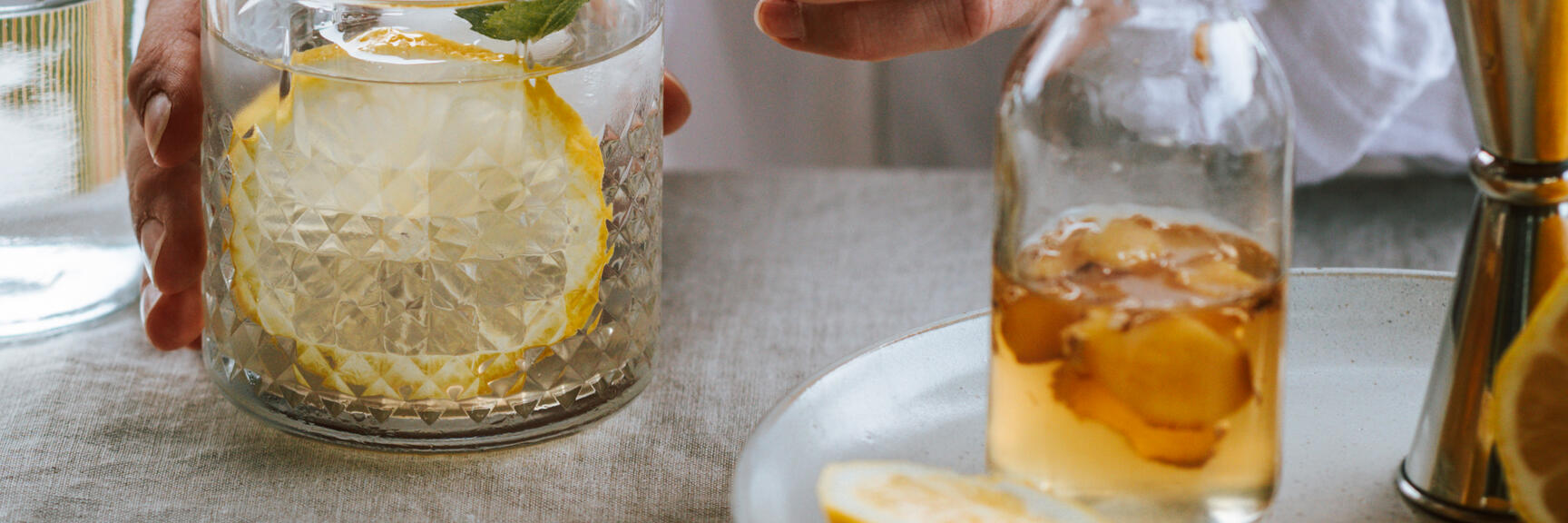 Hand garnishing cocktail glass with mint and lemon slice, syrup bottle and jigger beside on linen cloth.
