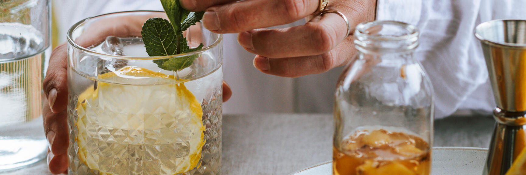 Hand garnishing cocktail glass with mint and lemon slice, syrup bottle and jigger beside on linen cloth.