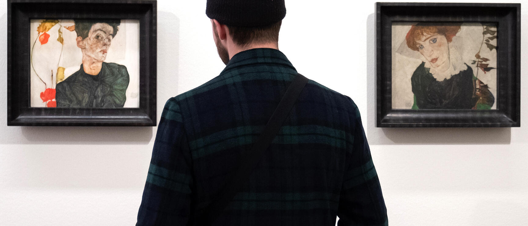 Man in cap viewing two expressionist paintings on white museum wall.