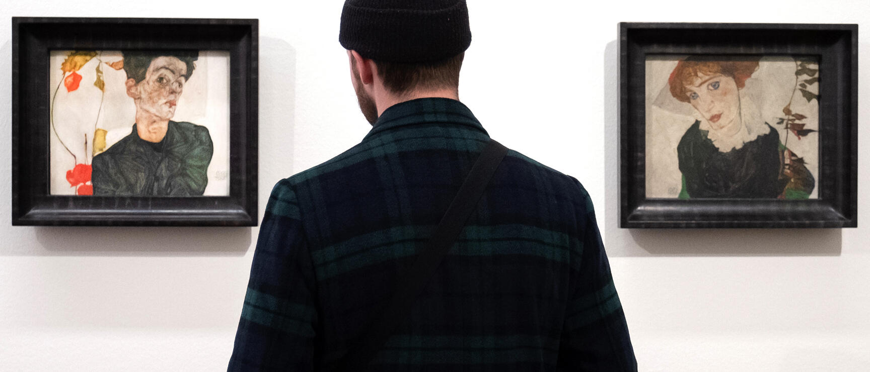 Man in cap viewing two expressionist paintings on white museum wall.