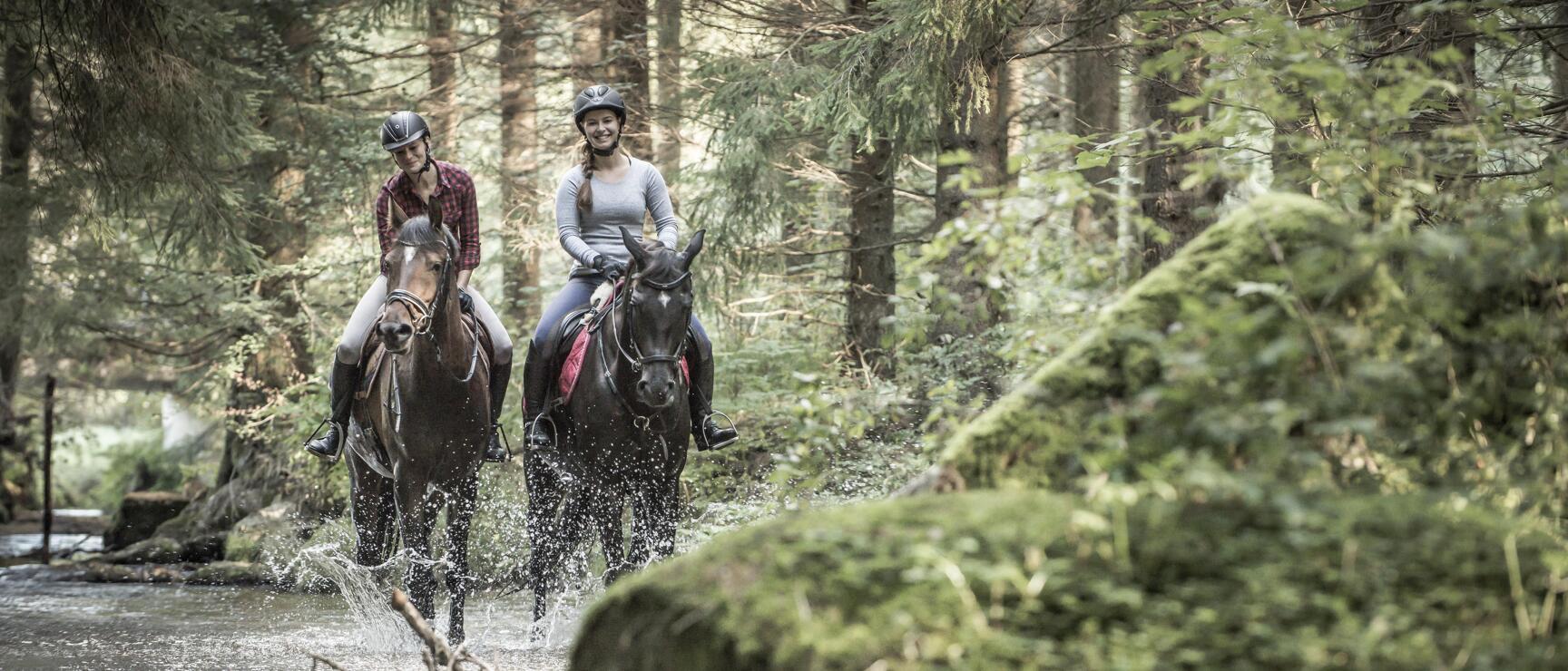 Two female horse riders with helmets crossing a stream on horseback in the forest, water splashing visible.