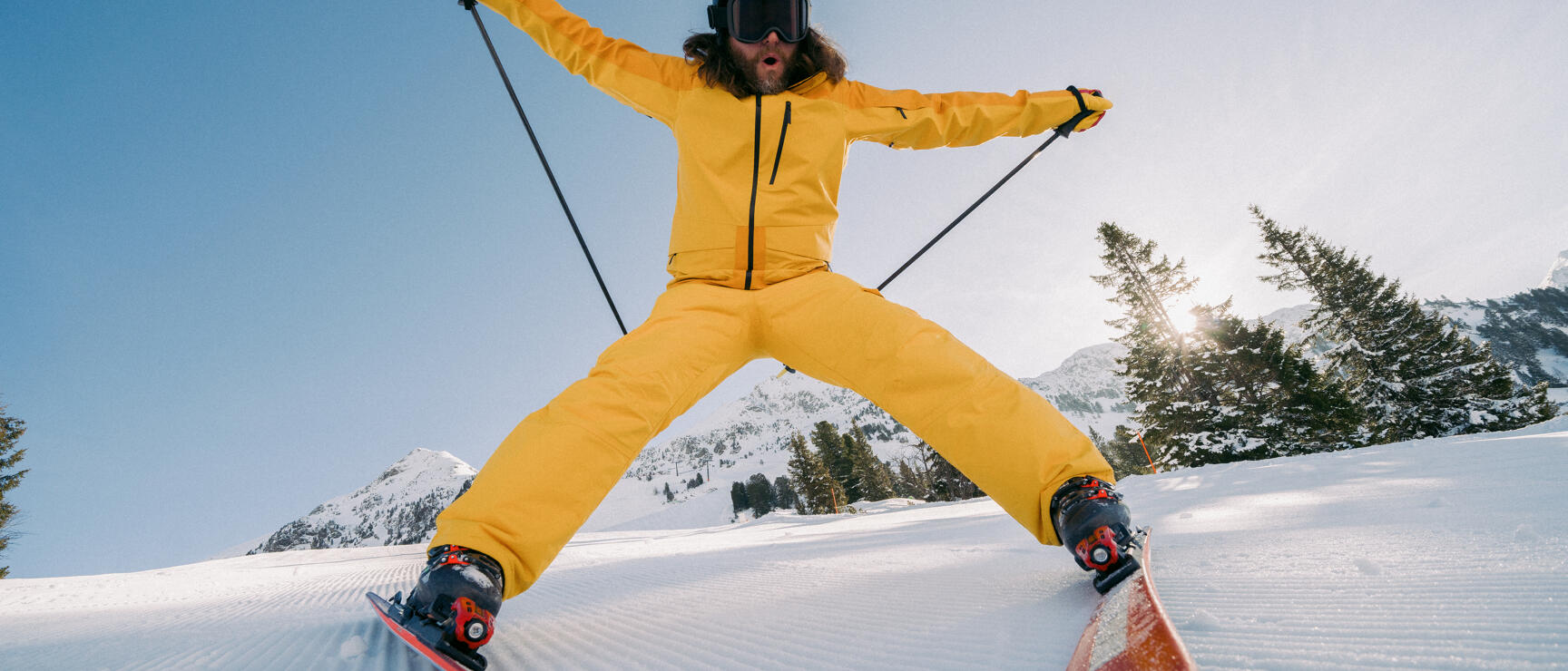 Skier in yellow suit jumping on groomed slope with snow-covered mountains and trees in background.