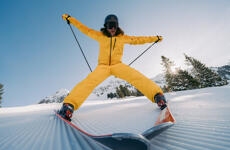 Skier in yellow suit jumping on groomed slope with snow-covered mountains and trees in background.