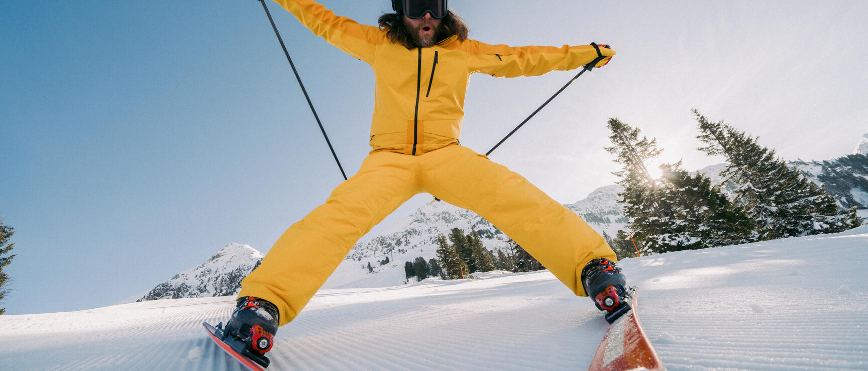 Skier in yellow suit jumping on groomed slope with snow-covered mountains and trees in background.