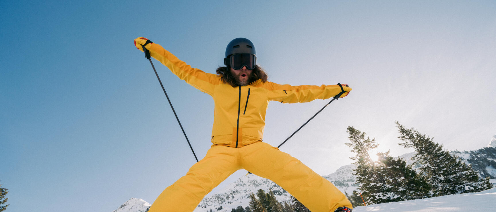 Skier in yellow suit jumping on groomed slope with snow-covered mountains and trees in background.