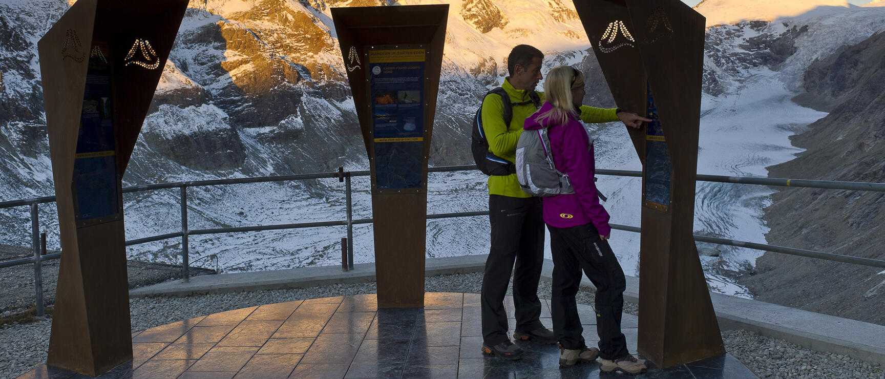 Two people at information panels on observation platform with Großglockner and Pasterze glacier in background.