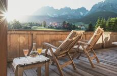 Wooden terrace with two deckchairs and drinks, view of mountain panorama and village in sunlight.
