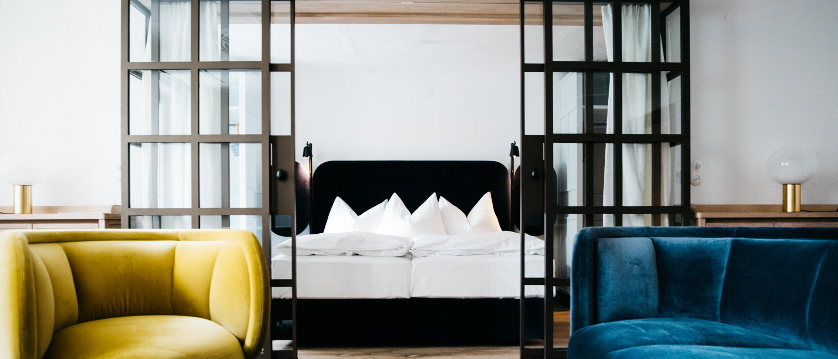 Hotel room with yellow armchair, blue sofa and double bed behind black glass partition.